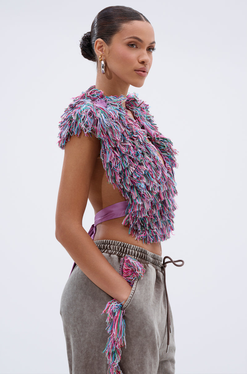 A woman with slicked-back hair confidently poses against a plain light background, wearing THE ONE FRINGE BLOUSE—a multicolor fringe yarn top—paired with loose gray drawstring loungewear pants.
