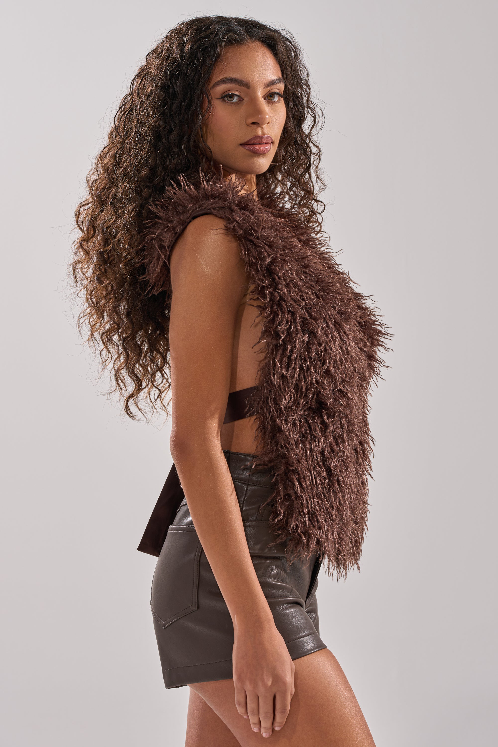 A woman with long curly hair wears the luxe, sleeveless HANNAH FUR BLOUSE and dark brown leather shorts, standing in profile against a light background—perfect for effortless luxury layering.