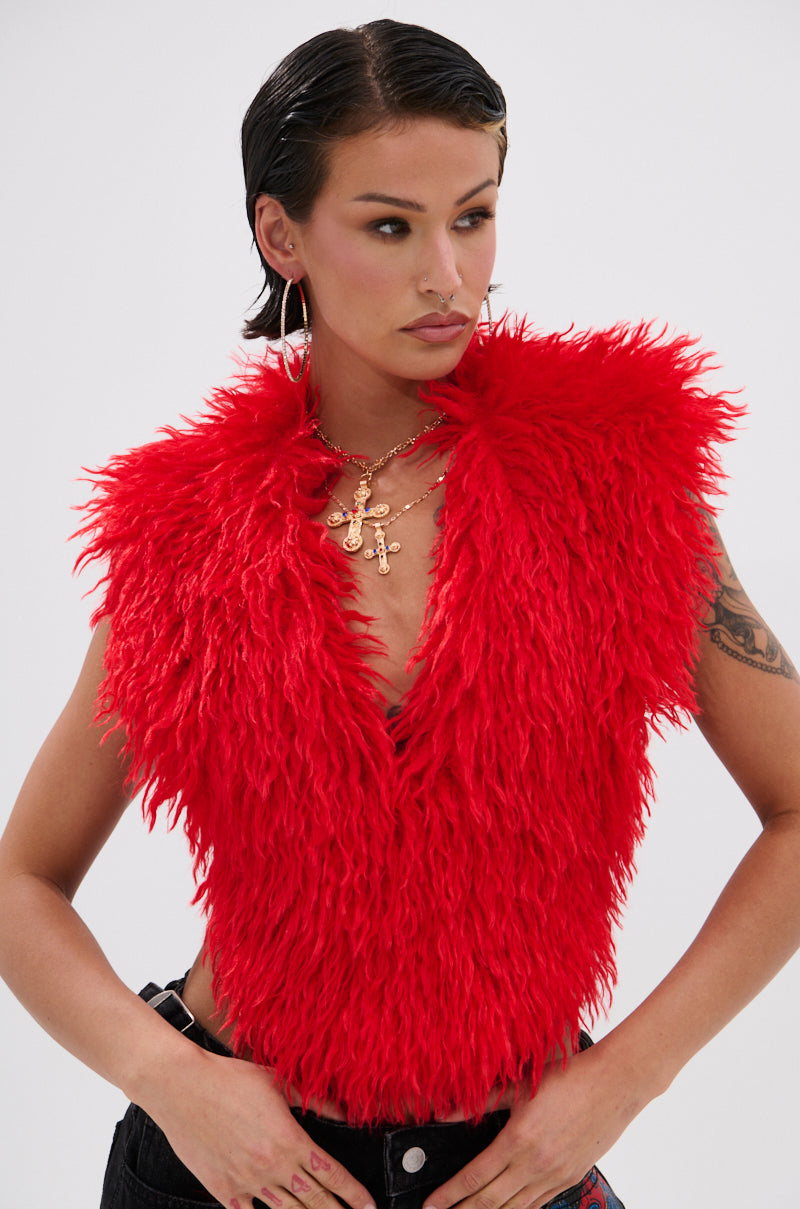 Wearing the CARLA FAUX FUR HALTER TOP in bright red, paired with gold cross pendant necklaces, hoop earrings, and black pants, a person stands against a plain light background with arms slightly bent.