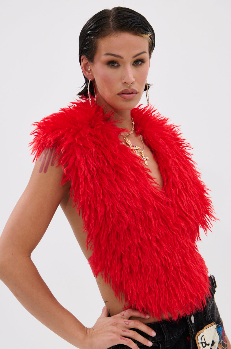 A person with short, slicked-back hair models the CARLA FAUX FUR HALTER TOP in bright red—a shaggy, cropped, sleeveless piece—styled with large hoop earrings and layered gold necklaces. They pose confidently with one hand on their hip against a plain background.