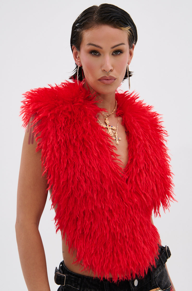A person with short dark hair wears the CARLA FAUX FUR HALTER TOP—a fluffy red faux fur piece with a plunging V-neckline—styled with gold layered necklaces, hoop earrings, and dark pants against a plain light background.