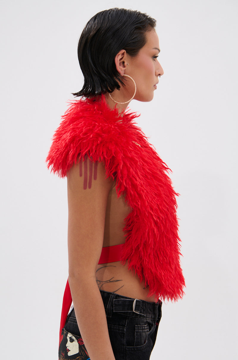 A person with short, wet-look dark hair and large hoop earrings wears the bright red CARLA FAUX FUR HALTER TOP in a cropped, sleeveless style with black jeans featuring a graphic patch, standing in profile against a plain background.
