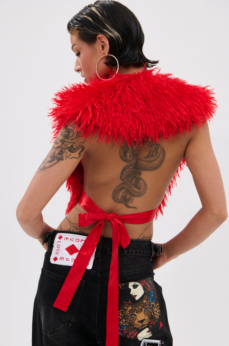 A person with wet, slicked-back hair wears the CARLA FAUX FUR HALTER TOP in bright red, featuring a plunging V-neckline and ribbon ties. They style it with large hoop earrings, visible tattoos, and black jeans with colorful patches and cards showing.