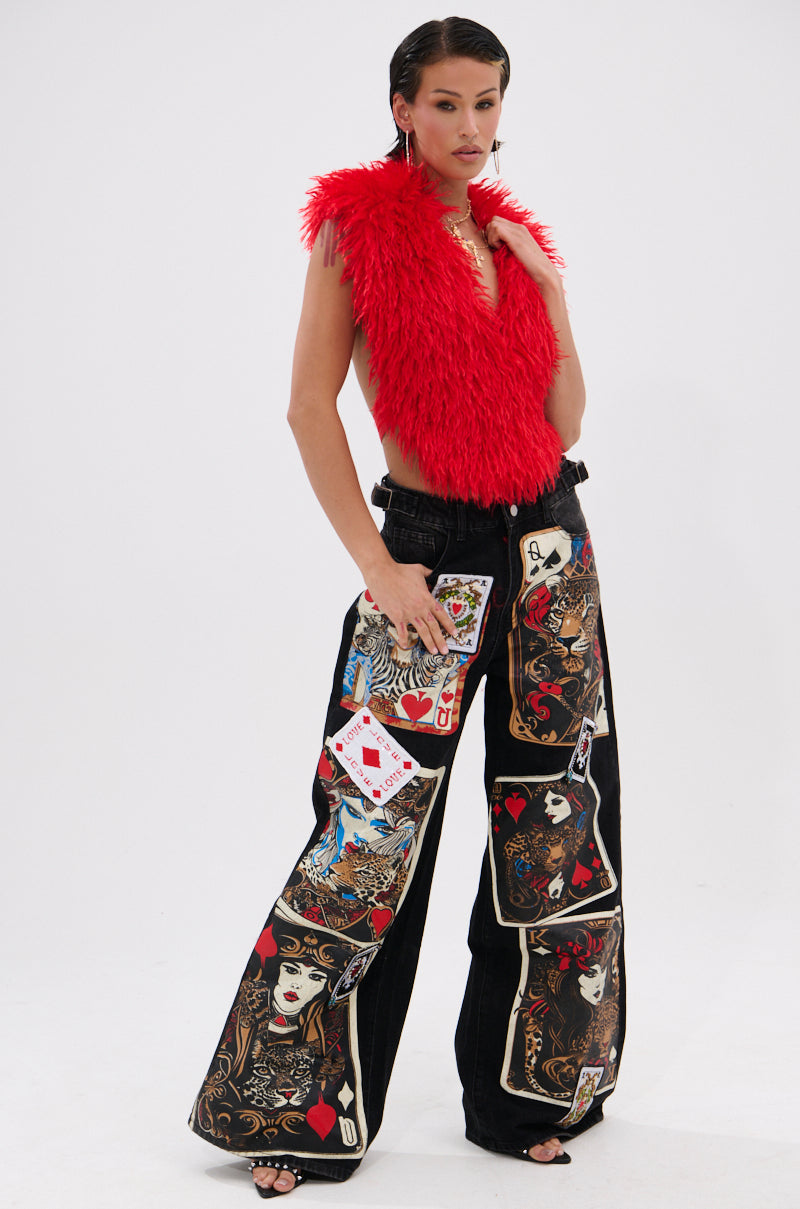 A person stands against a plain white background, wearing the bright red CARLA FAUX FUR HALTER TOP with a cropped sleeveless silhouette and wide-leg black pants featuring bold, colorful playing card designs.
