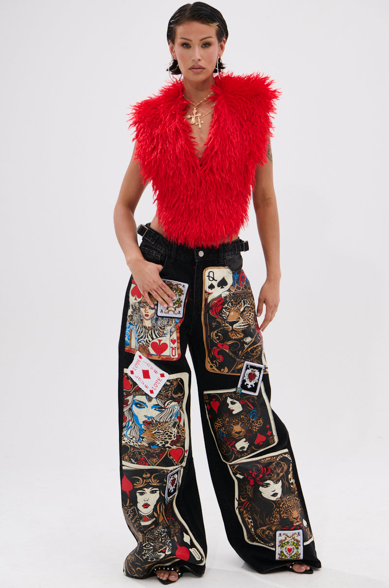 A person wears the CARLA FAUX FUR HALTER TOP in bright red, styled with wide-leg black pants featuring bold playing card and artistic face prints, against a plain white background.