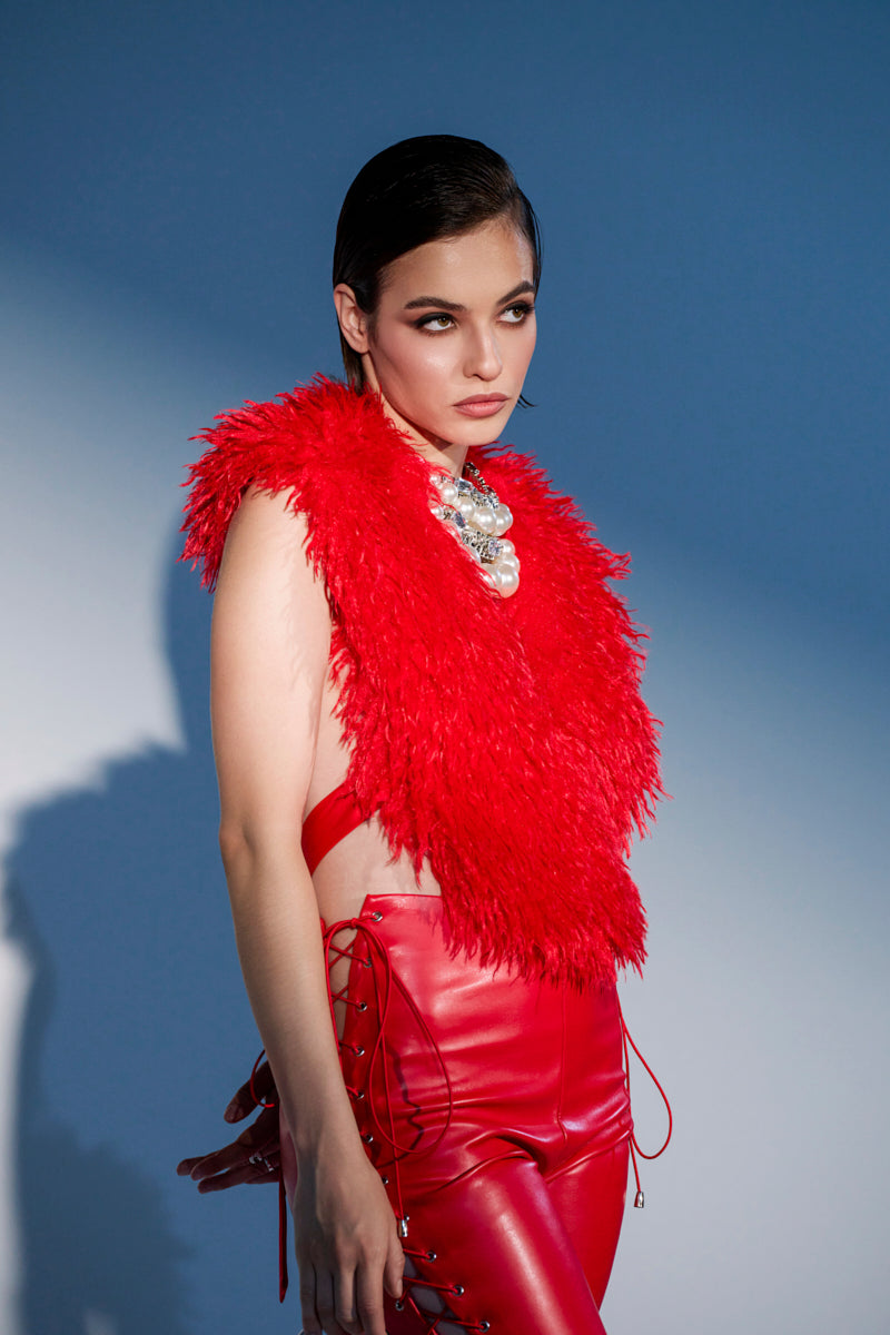 A woman with dark hair wears the bold, cropped CARLA FAUX FUR HALTER TOP in red and matching high-waisted red leather pants with lace-up sides, accessorized with a chunky metallic necklace, against a blue background.