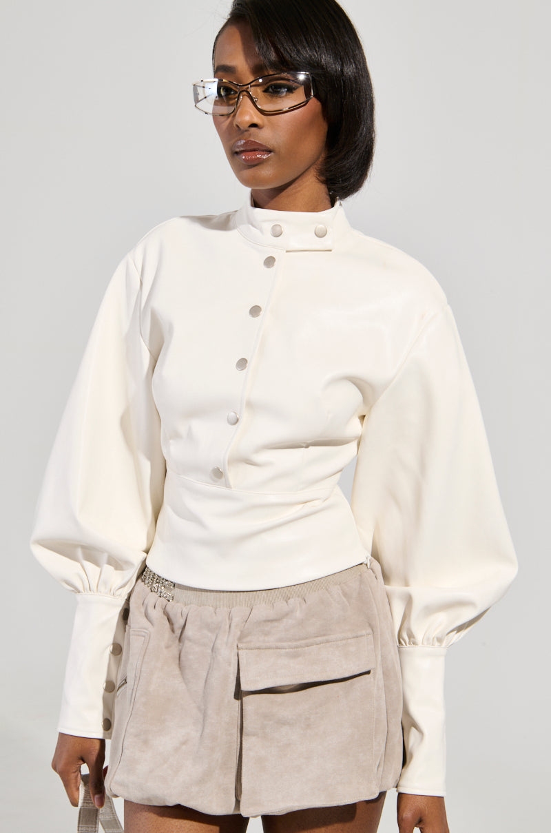 A person wearing a stylish outfit poses against a neutral background. They are dressed in the NOT YOUR NORMAL BIKER CHICK TOP, a white buttoned blouse with puffed sleeves and an edgy mock neck collar, paired with a beige skirt featuring large pockets. To complete the look, they wear rectangular glasses and maintain a neutral expression.