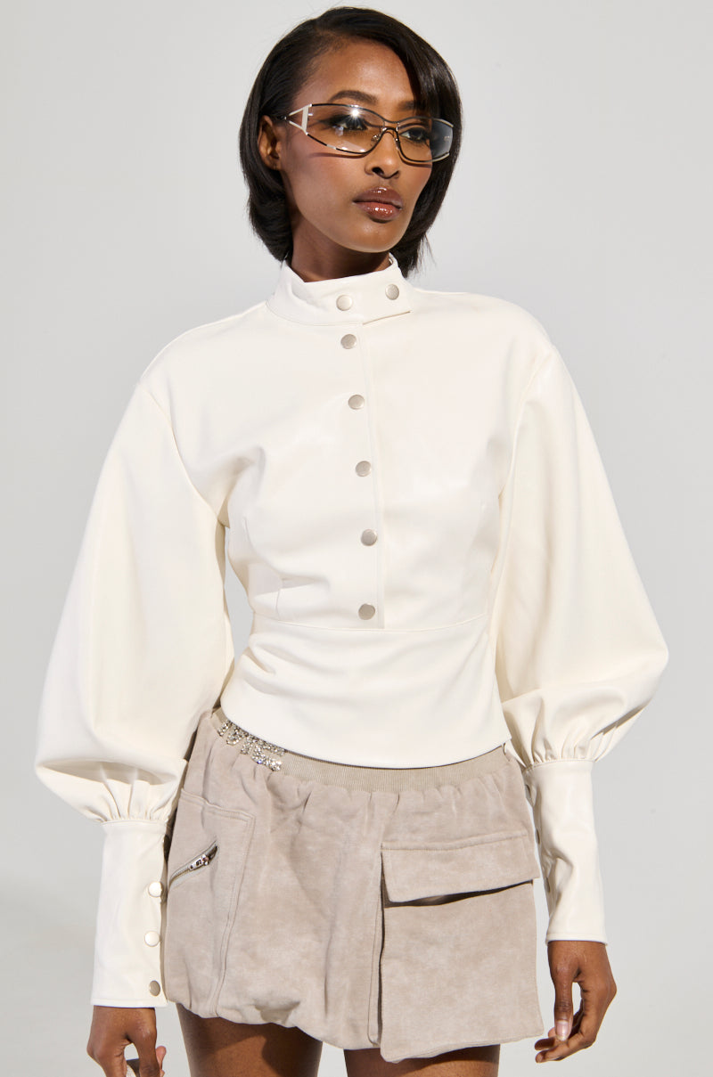 A person wearing a stylish ensemble featuring the "NOT YOUR NORMAL BIKER CHICK TOP," a white buttoned, long-sleeve shirt with puffed shoulders, paired with a beige suede skirt with pockets. The outfit is given an edgy vibe with rectangular glasses and a faux leather jacket, set against a neutral background.