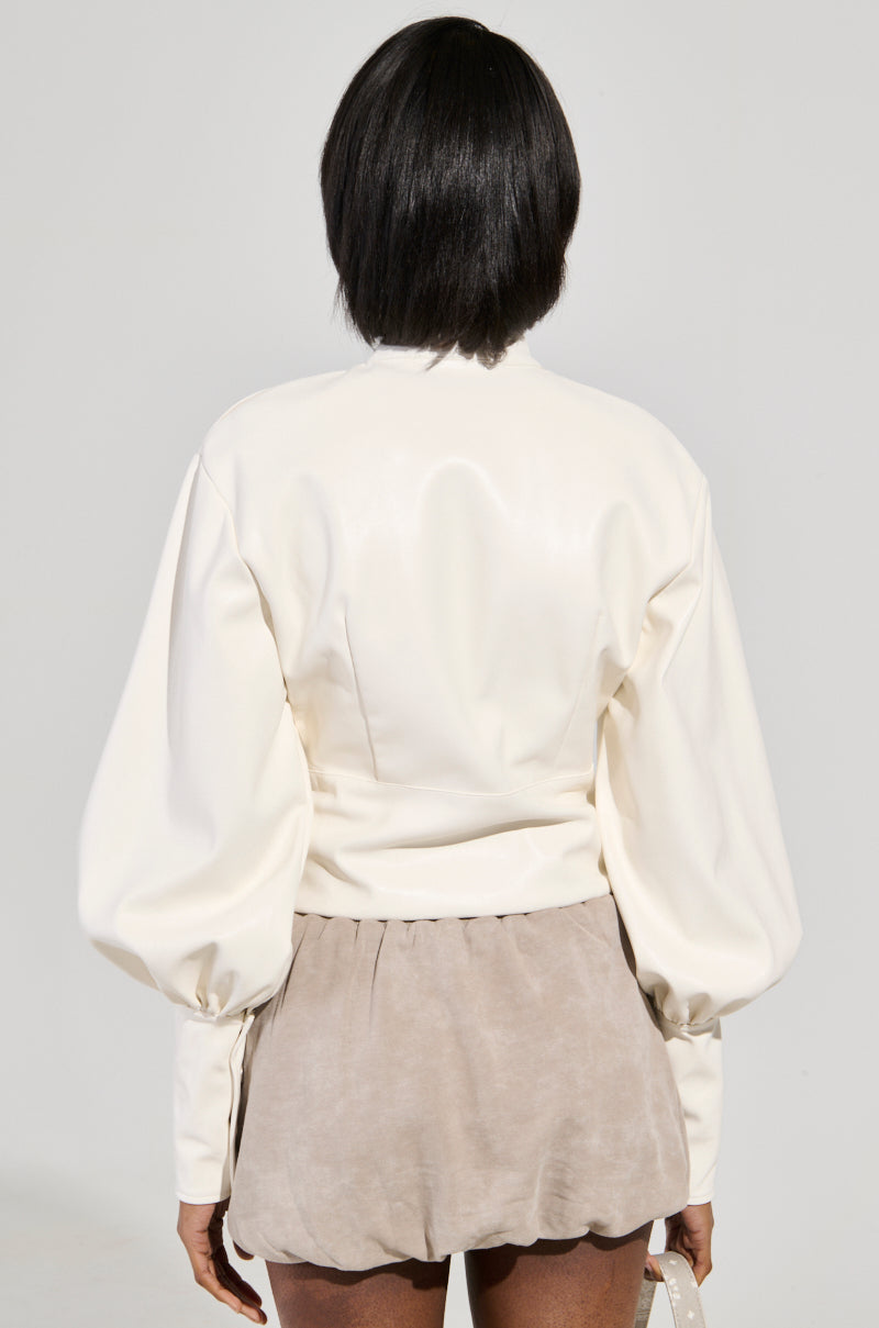 A person with short, dark hair is standing with their back to the camera, dressed in the NOT YOUR NORMAL BIKER CHICK TOP— a white long-sleeved top featuring puffy sleeves and a mock neck collar—paired with light brown shorts. The plain, light-colored background adds a subtle touch.
