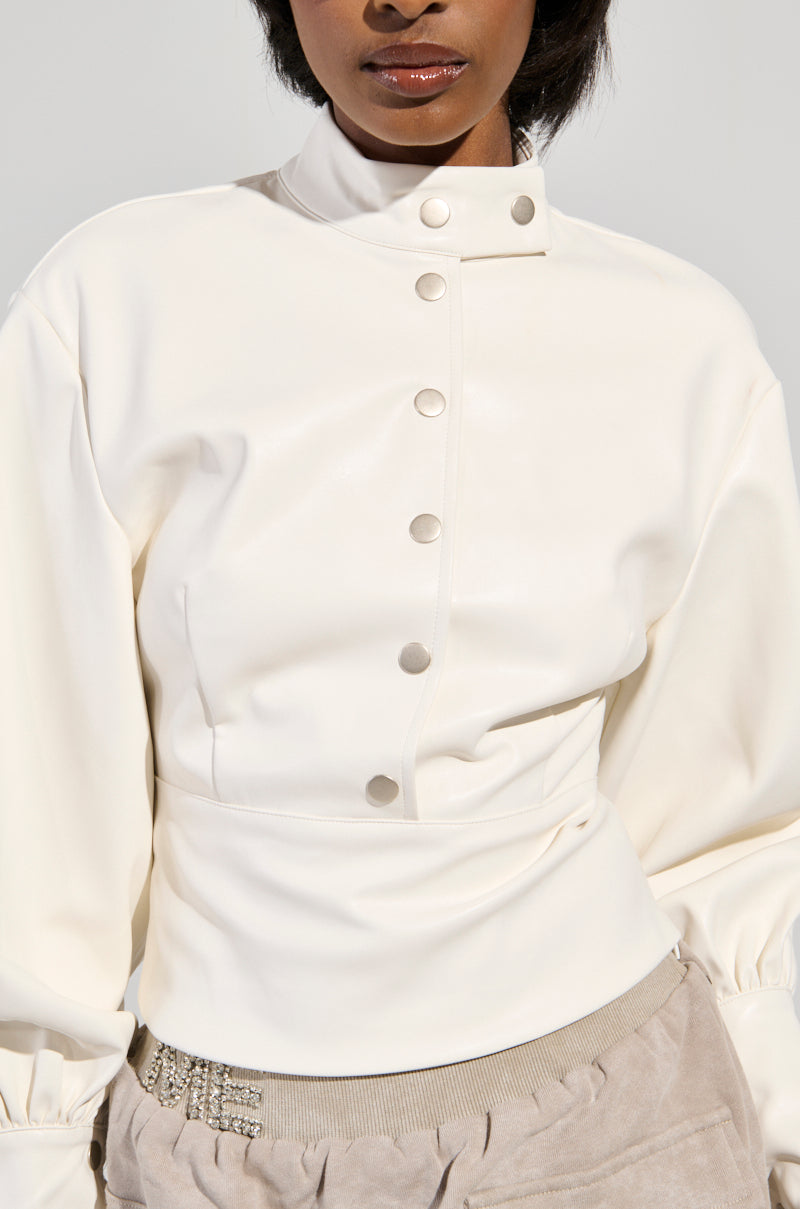 A person is featured from chest to chin wearing the NOT YOUR NORMAL BIKER CHICK TOP, a high-neck, long-sleeve white button-up with metallic buttons. The mock neck collar provides a sleek touch, and the ensemble is paired with taupe pants featuring silver ornate detailing on the waistband.