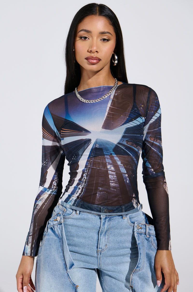 A city-slicker with long dark hair wears the "SKYS THE LIMIT" mesh long sleeve top, featuring a geometric design, and pairs it with light blue jeans. She accessorizes with a silver chain necklace and hoop earrings, posing confidently against a plain white background.