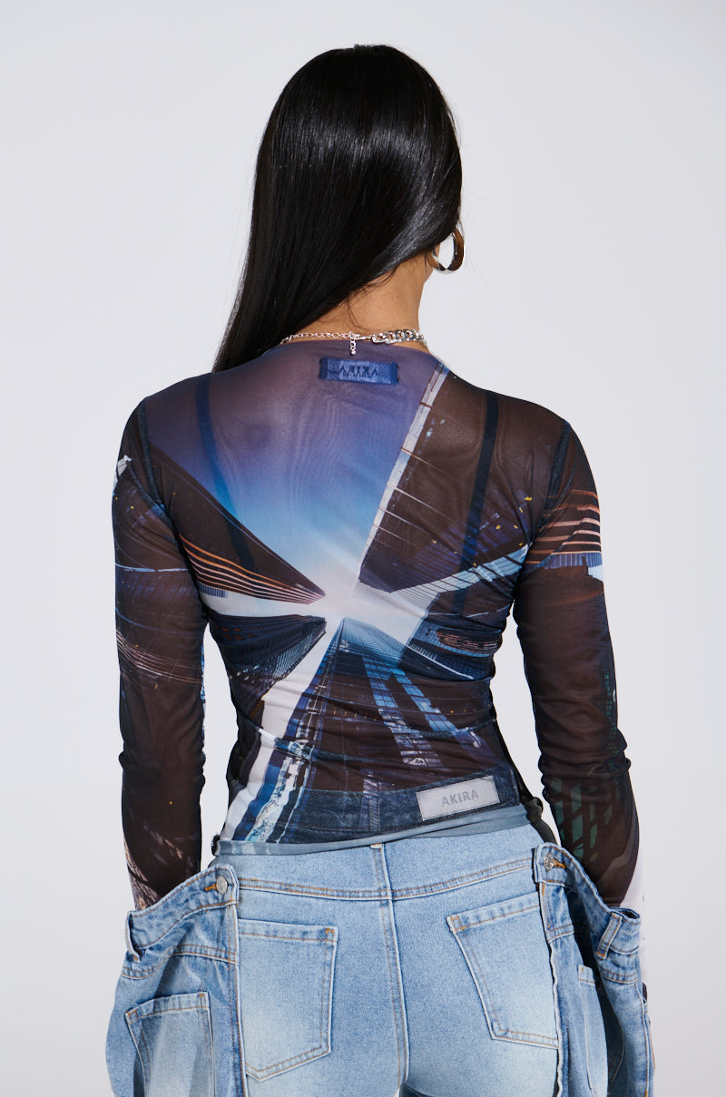 A city-slicker with long dark hair wears the SKYS THE LIMIT MESH LONG SLEEVE TOP featuring a skyscraper graphic, paired with light blue jeans. They stand against a plain background, highlighting the top's design and the jeans' fit.