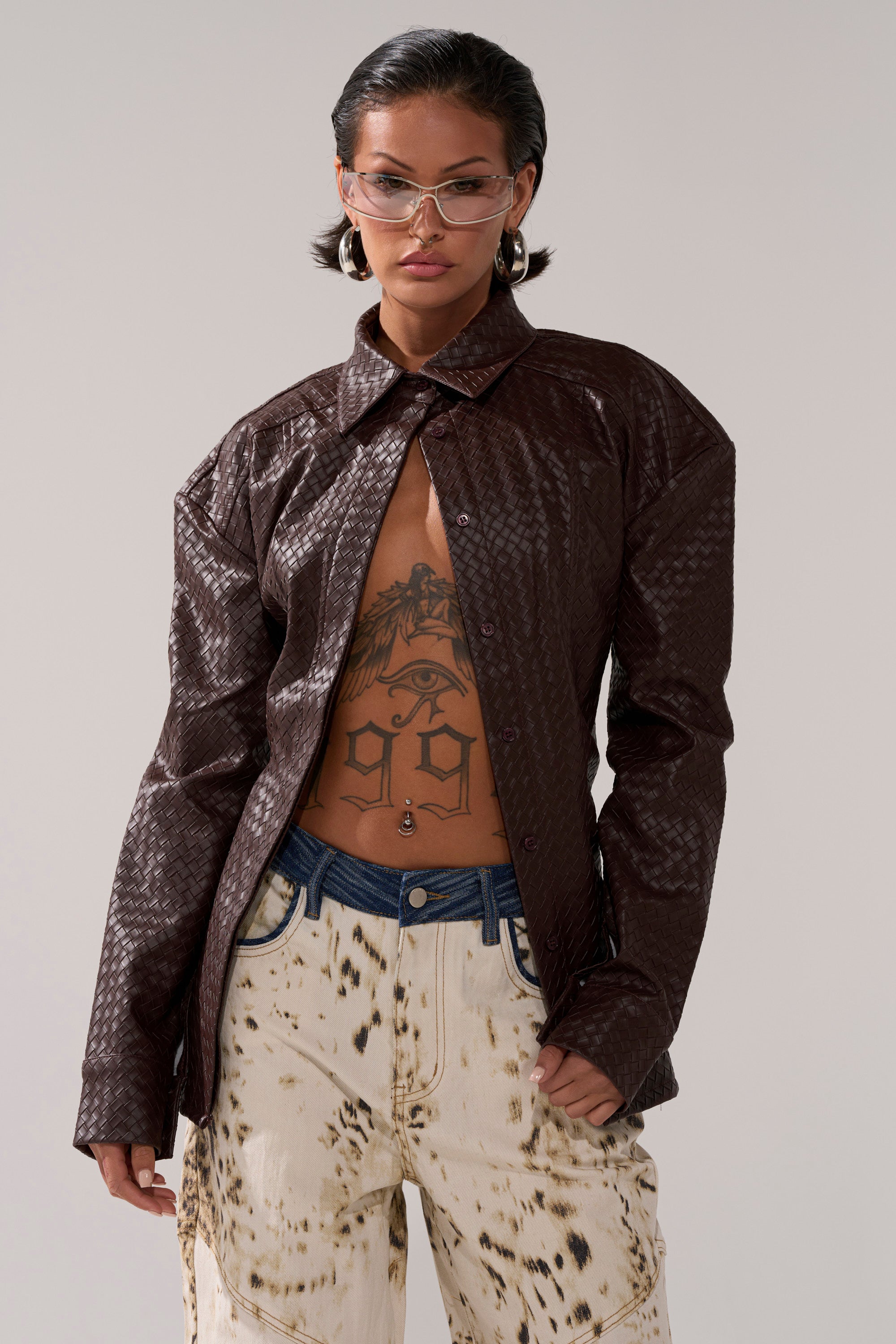 A person with short dark hair, clear glasses, and a chest tattoo wears the FEELING RICH FAUX LEATHER BUTTON DOWN left open over paint-splattered jeans, posing confidently against a plain background.