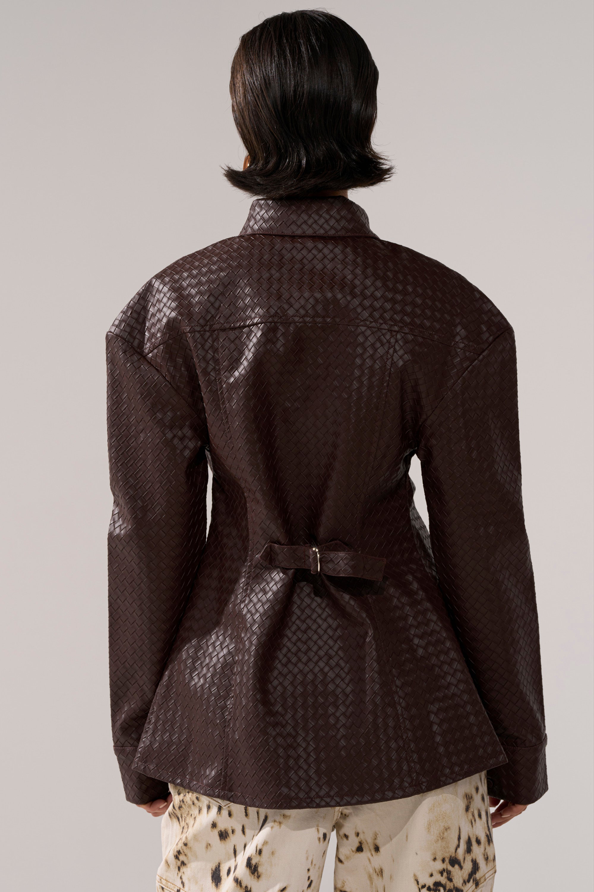 A person with short, dark hair is seen from behind wearing the FEELING RICH FAUX LEATHER BUTTON DOWN and cream-colored pants with dark abstract spots.