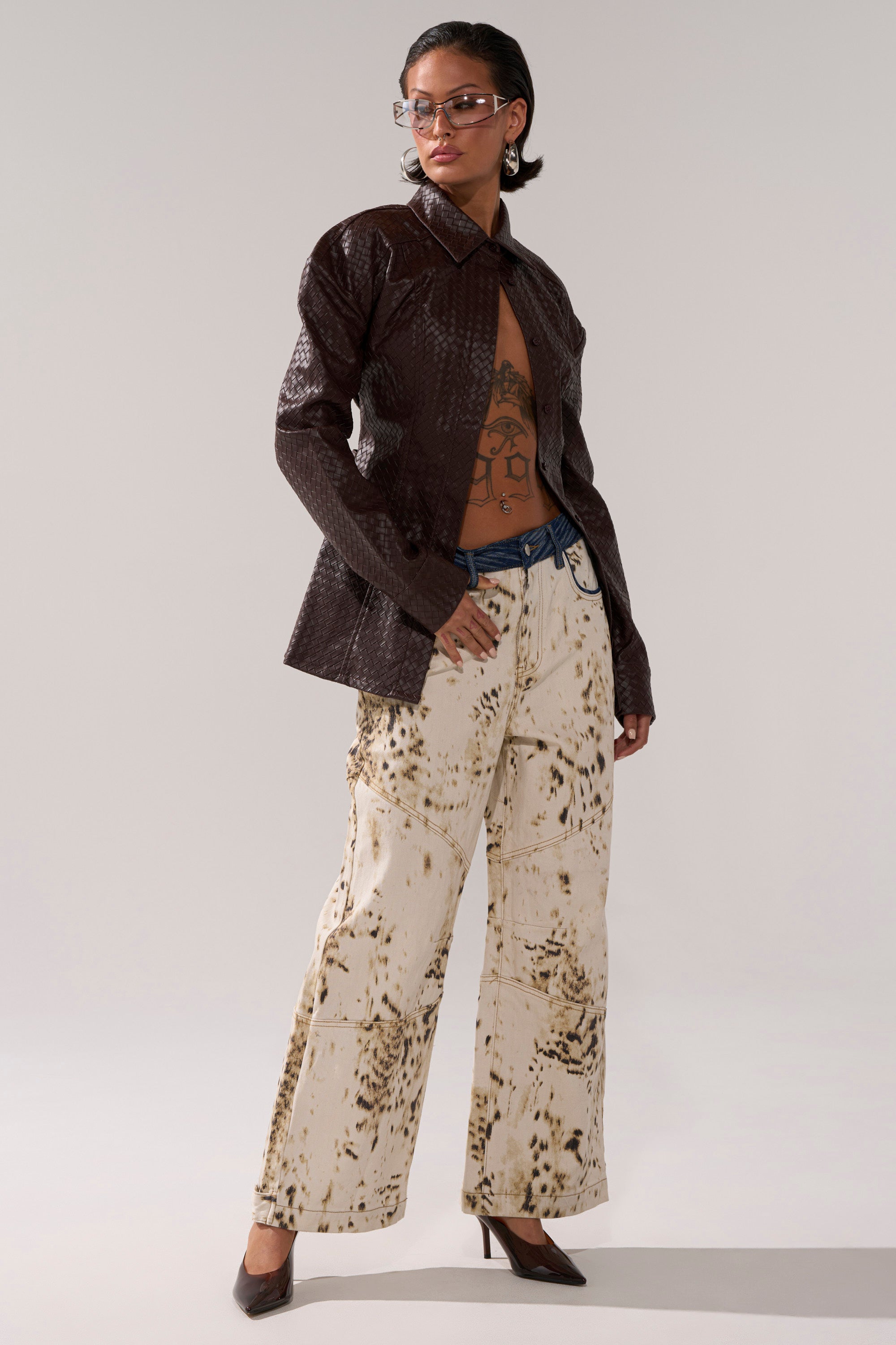 A person with short hair and glasses wears the FEELING RICH FAUX LEATHER BUTTON DOWN, left mostly open to reveal a tattooed chest, paired with high-waisted cream wide-leg pants with a brown splatter pattern and brown heels.