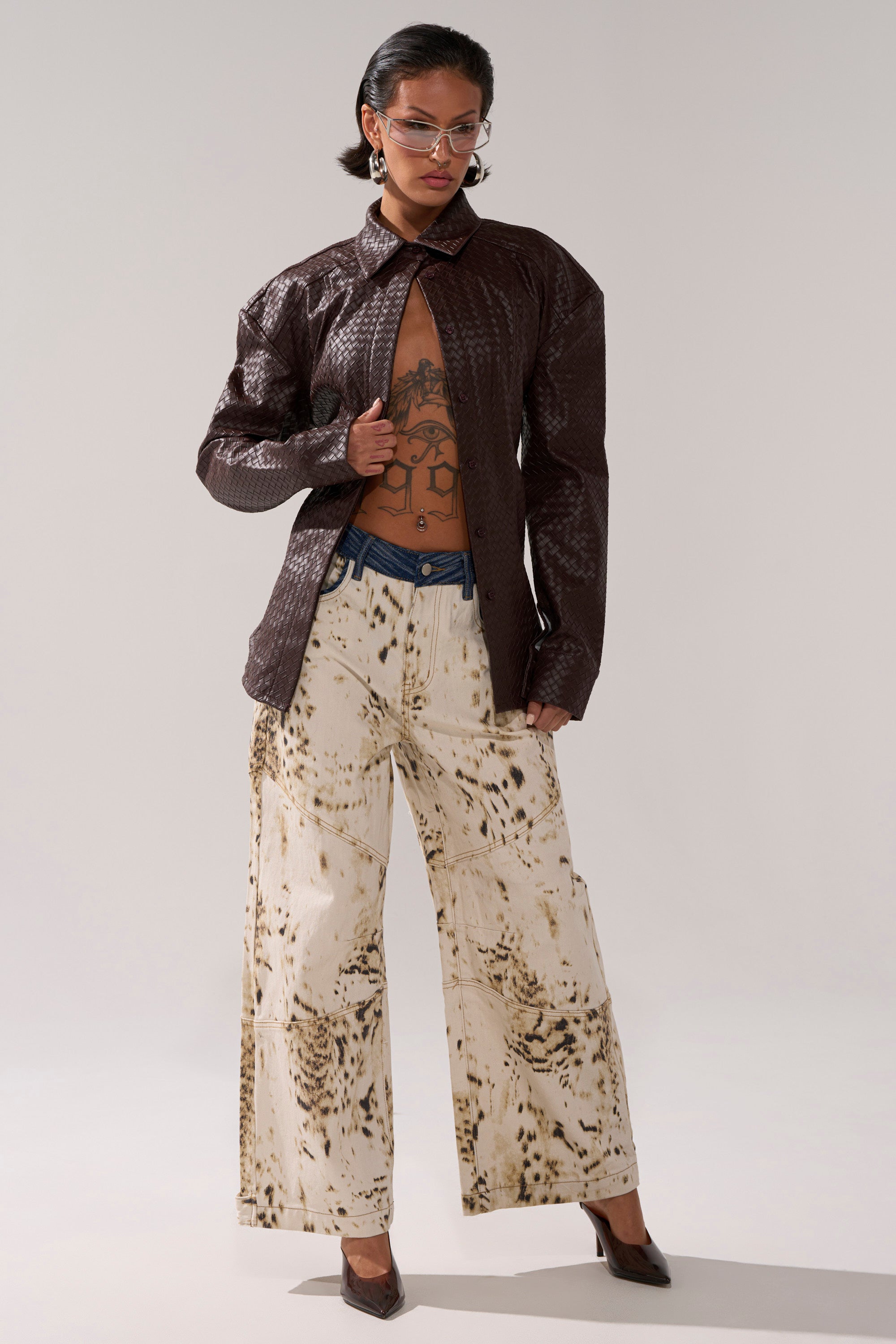 A person stands confidently in the FEELING RICH FAUX LEATHER BUTTON DOWN, open to reveal a tattooed chest, paired with cream wide-leg pants with a brown abstract pattern, a blue waistband detail, an adjustable buckled belt, clear glasses, and dark pointed heels.