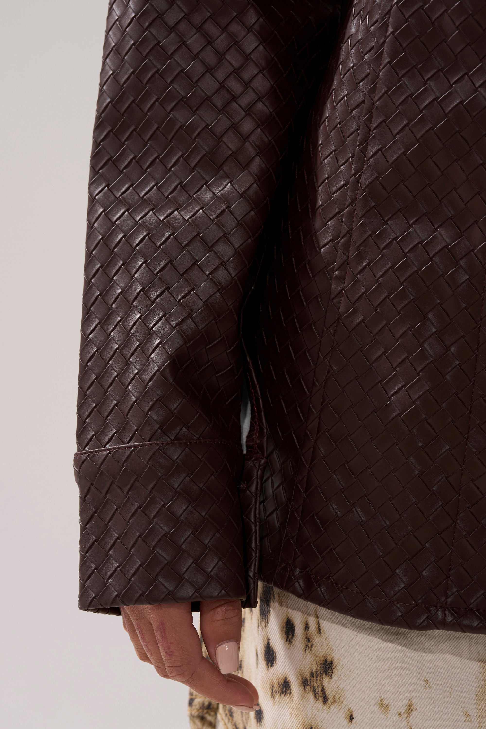 A close-up of a person wearing the FEELING RICH FAUX LEATHER BUTTON DOWN, featuring dark brown woven faux leather and contour paneling, with their hand by their side and a light, spotted fabric layered underneath.