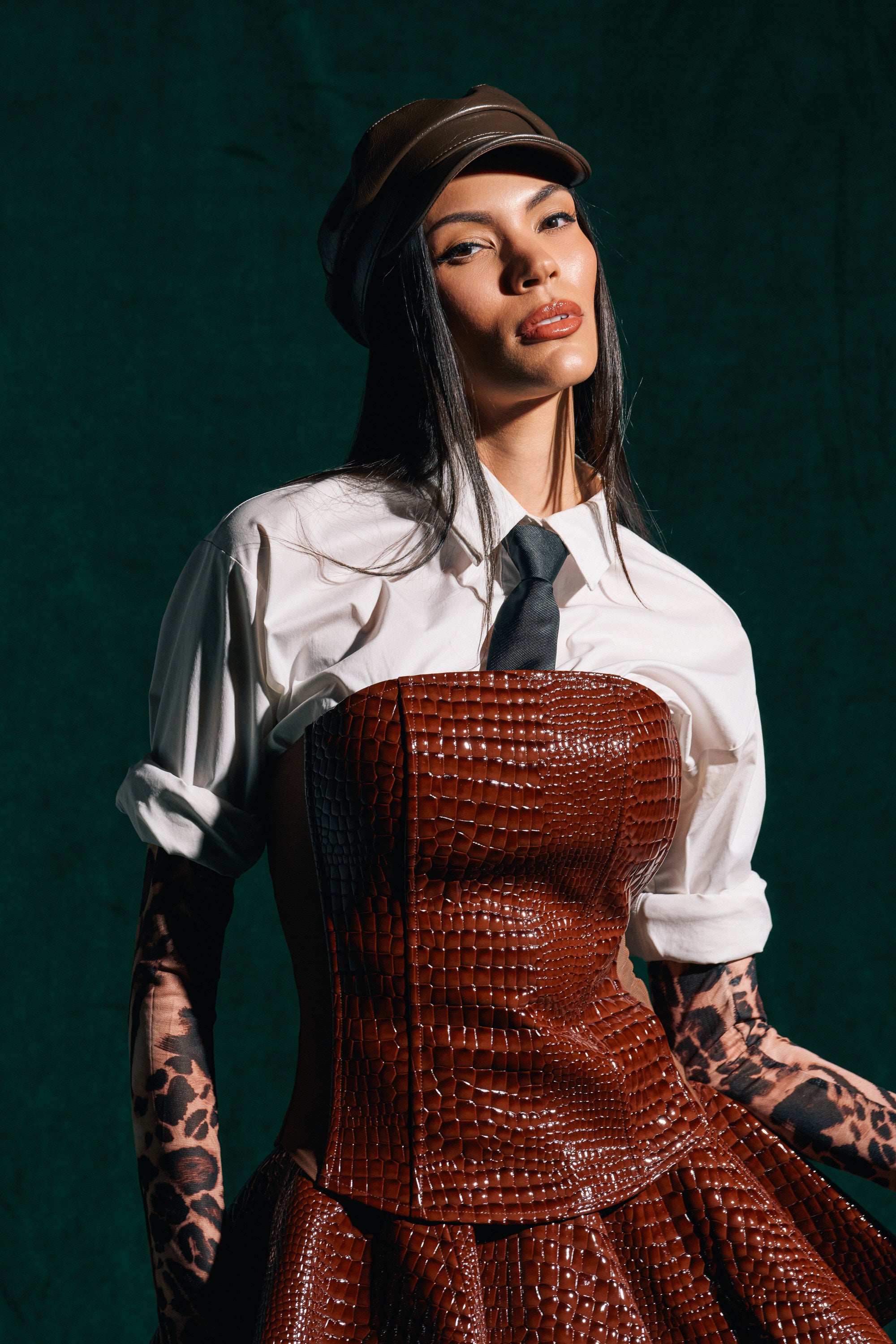 A person poses confidently against a dark green background wearing a white shirt, black tie, brown leather cap, leopard-print gloves, and the TOO GOOD FOR YOU CROCODILE CORSET.