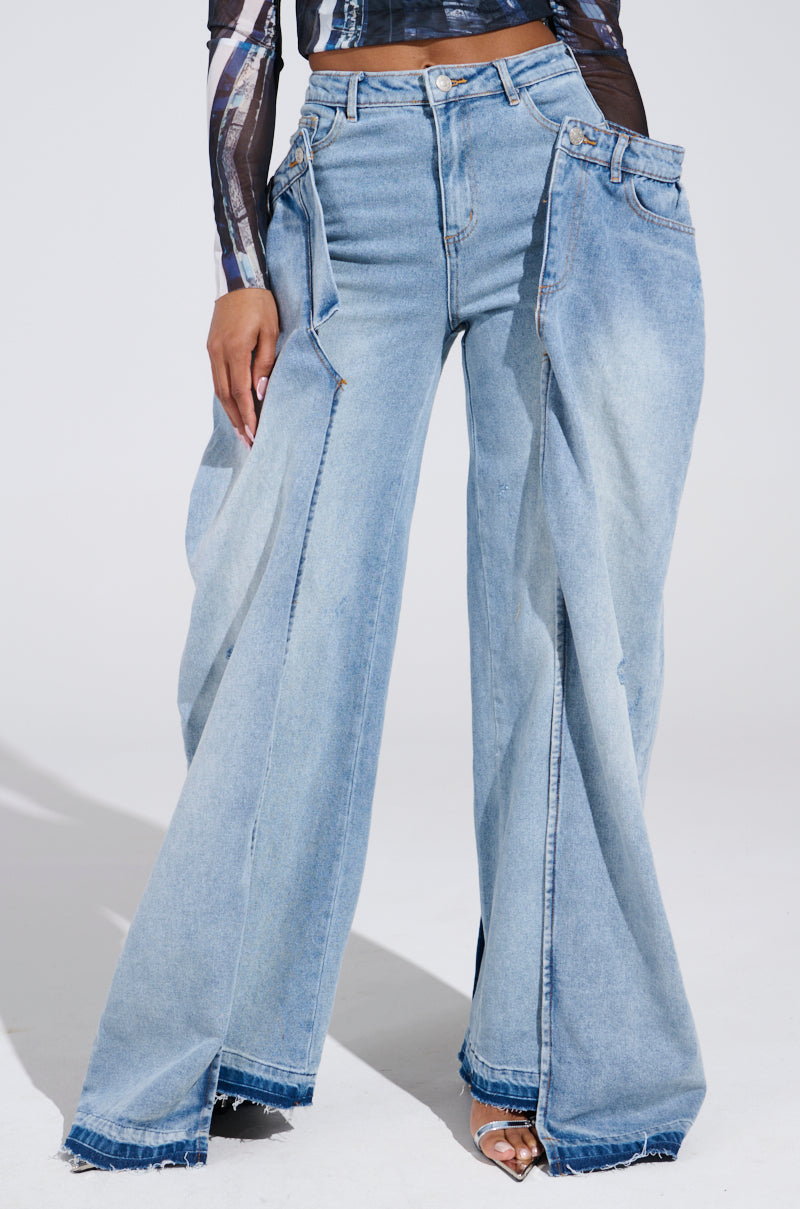 A person wears SEEING DOUBLE DECONSTRUCTED DENIM JEANS, characterized by high-waisted, wide-leg design with overlapping layers and frayed hems, paired with a black and white long-sleeved top. The studio photo has a neutral background.