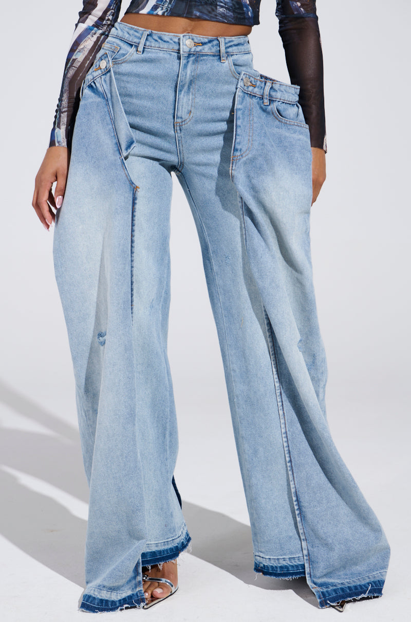 A person flaunts SEEING DOUBLE DECONSTRUCTED DENIM JEANS, featuring a high-rise fit with double-layered wide-leg denim and an overlapping design. The light blue jeans, with exaggerated flares and layered waistbands, perfectly complement a dark long-sleeve top and open-toe heels.