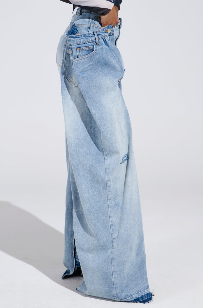 A person is wearing oversized SEEING DOUBLE DECONSTRUCTED DENIM JEANS in light blue, standing against a plain background. The high-rise fit accentuates the waist, and the long length slightly covers the shoes. Hands are casually placed in pockets, adding to the laid-back vibe.