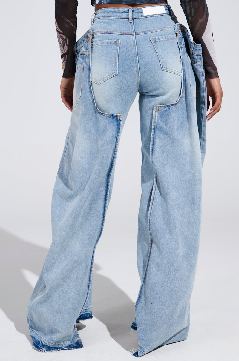 The person is wearing SEEING DOUBLE DECONSTRUCTED DENIM JEANS in light blue with a detachable panel design, visible seams, and back pockets. The jeans have a relaxed fit and are paired with a dark, long-sleeved top that partially shows their torso.