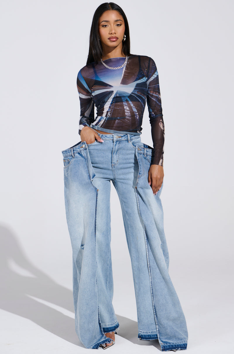A person wearing oversized SEEING DOUBLE DECONSTRUCTED DENIM JEANS with high-rise, wide-leg fit and multiple waistbands, paired with a fitted abstract-patterned long-sleeve top against a plain white background for a striking look.