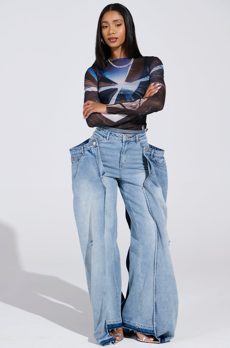 A woman stands against a plain backdrop wearing a sheer, patterned long-sleeve top and oversized SEEING DOUBLE DECONSTRUCTED DENIM JEANS with an exposed-pocket design. Her long, straight hair complements her open-toed shoes.