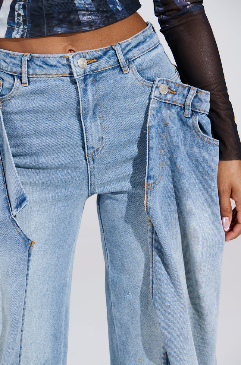 A person is seen wearing SEEING DOUBLE DECONSTRUCTED DENIM JEANS in statement blue with unique pocket details. The jeans complement a tucked-in black sheer long-sleeve top. The background is light gray, showing the midsection and part of one arm.