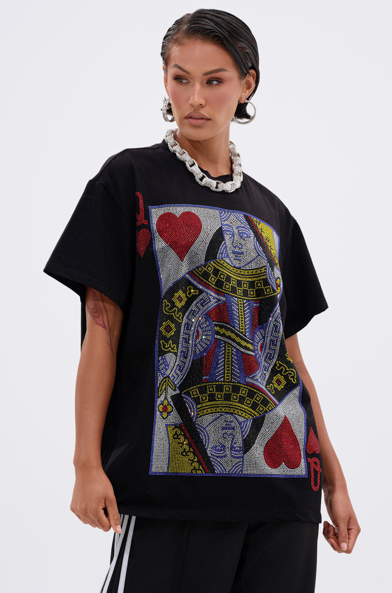 A woman in black pants and a chunky white necklace poses in THE REAL QUEEN OF HEARTS OVERSIZED TEE, which features a bold Queen of Hearts playing card graphic in red, yellow, and blue.