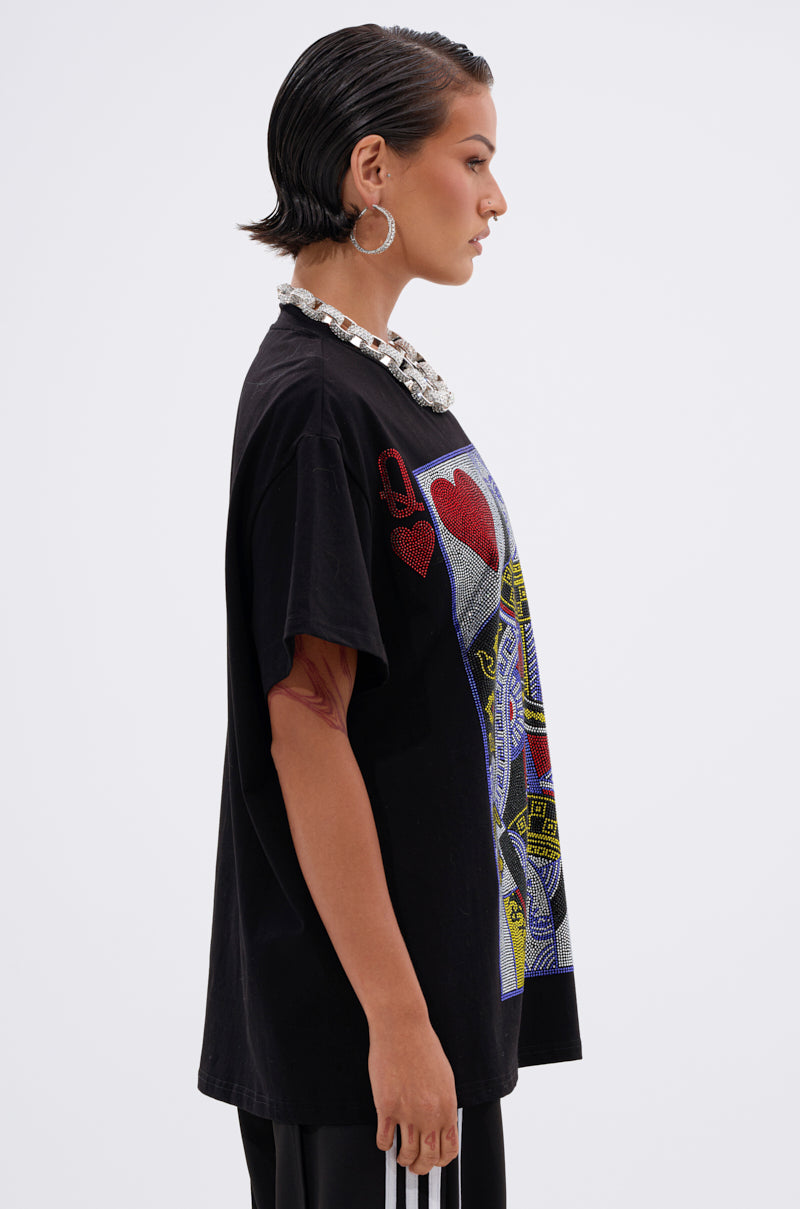 Woman in profile wears THE REAL QUEEN OF HEARTS OVERSIZED TEE with black pants featuring white stripes, a chunky silver necklace, and large hoop earrings, set against a plain white background.