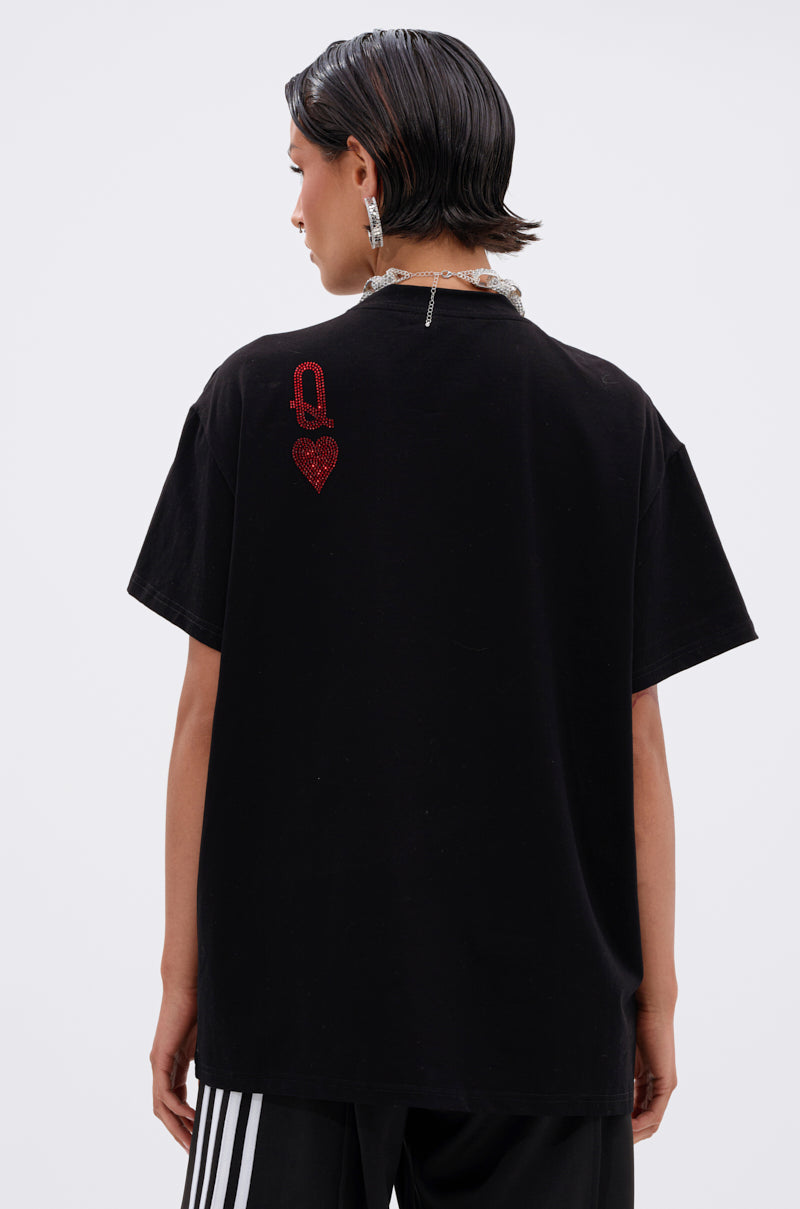 A person with short dark hair faces away from the camera, wearing THE REAL QUEEN OF HEARTS OVERSIZED TEE—a black graphic shirt with a small red "Q" and heart on the upper back—paired with black pants featuring white stripes.