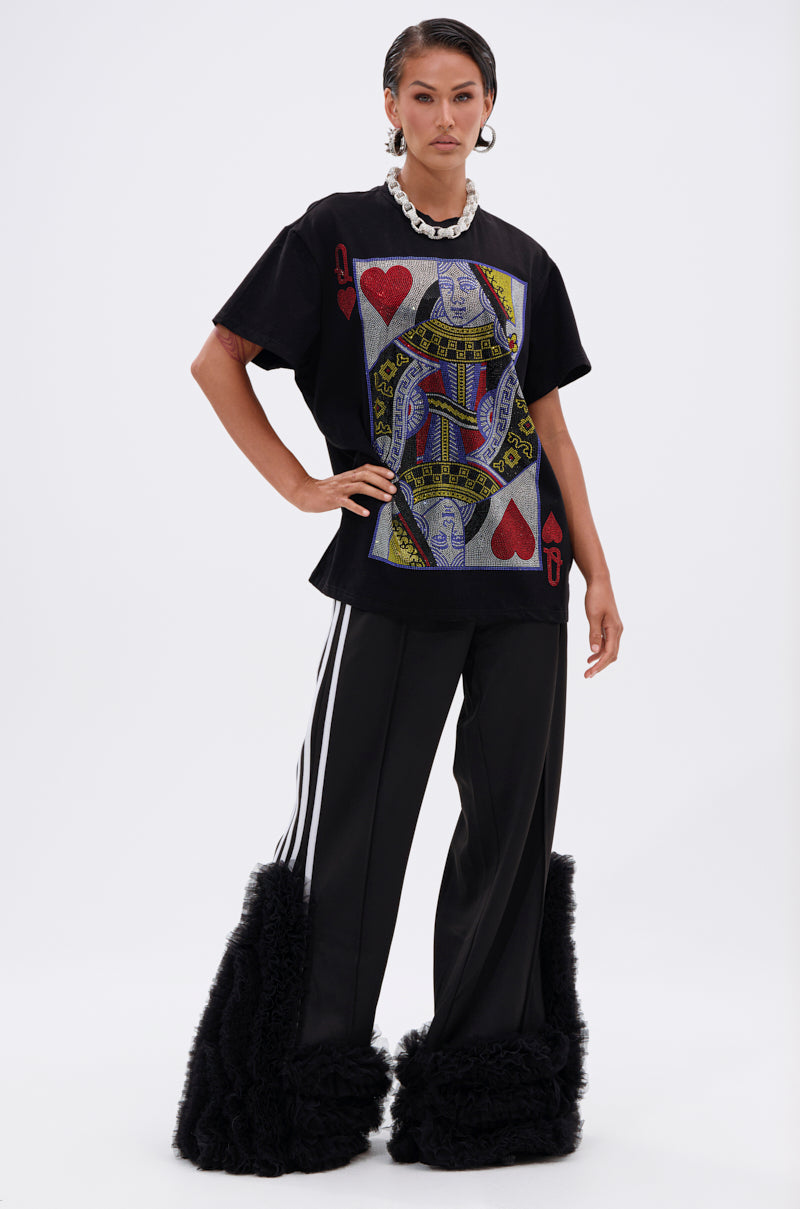 Wearing THE REAL QUEEN OF HEARTS OVERSIZED TEE featuring a bold playing card graphic, paired with black wide-leg pants with white stripes, black furry leg warmers, and a chunky white necklace.