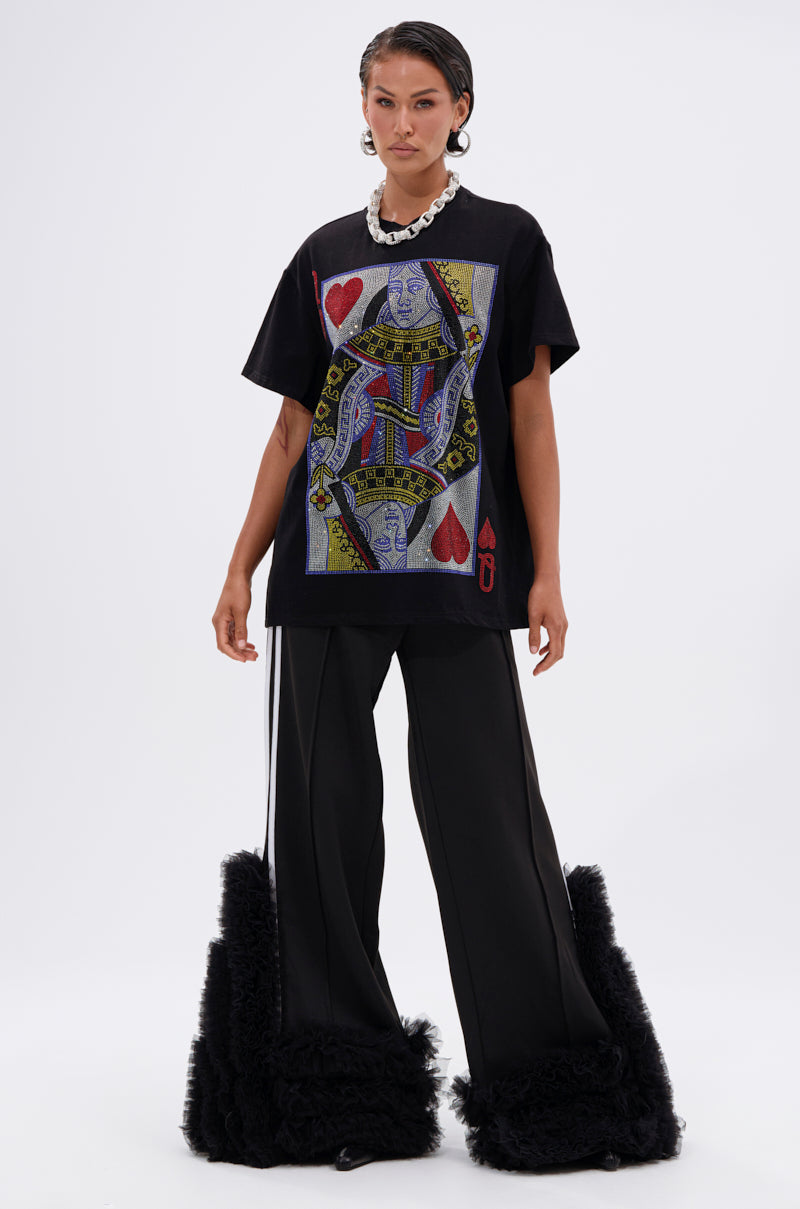 A person stands facing forward in THE REAL QUEEN OF HEARTS OVERSIZED TEE, featuring a bold playing card graphic, paired with wide-leg black pants with shaggy cuffs. The background is plain white.
