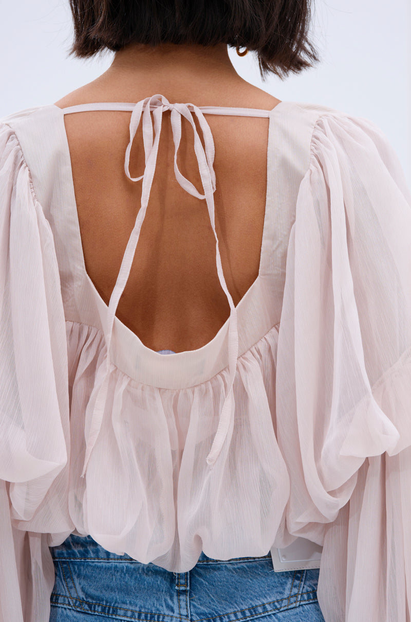 A woman with short, dark hair wears a sheer, flowy light pink SLOUCHY SQUARE NECK LONG SLEEVE BLOUSE TOP with billowy sleeves and an open back tied at the neck, paired effortlessly with blue denim jeans.
