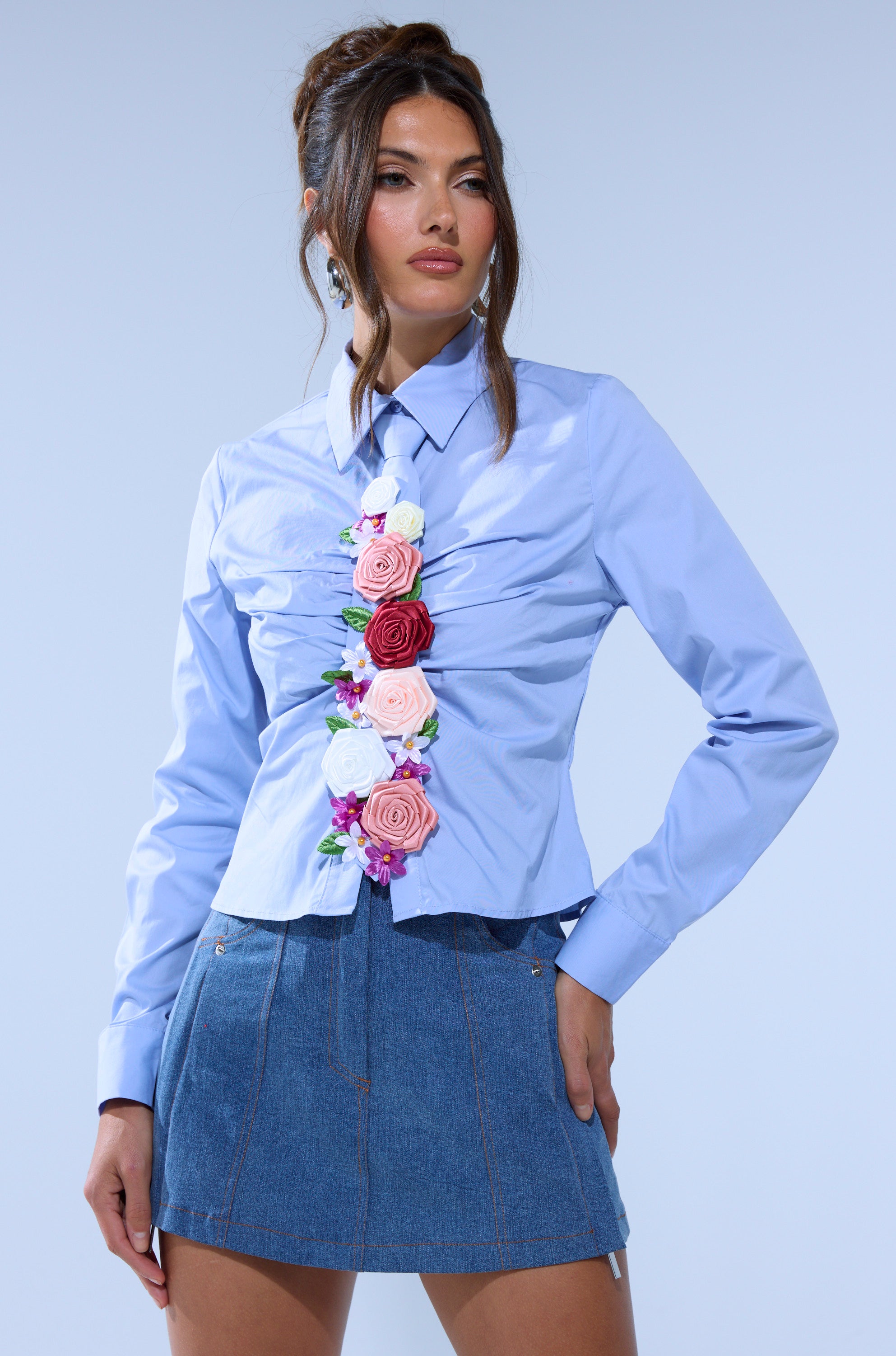 A woman wears a tailored fit BOUQUET BUTTON DOWN SHIRT with colorful floral appliques and a short denim skirt, posing against a plain light blue background.