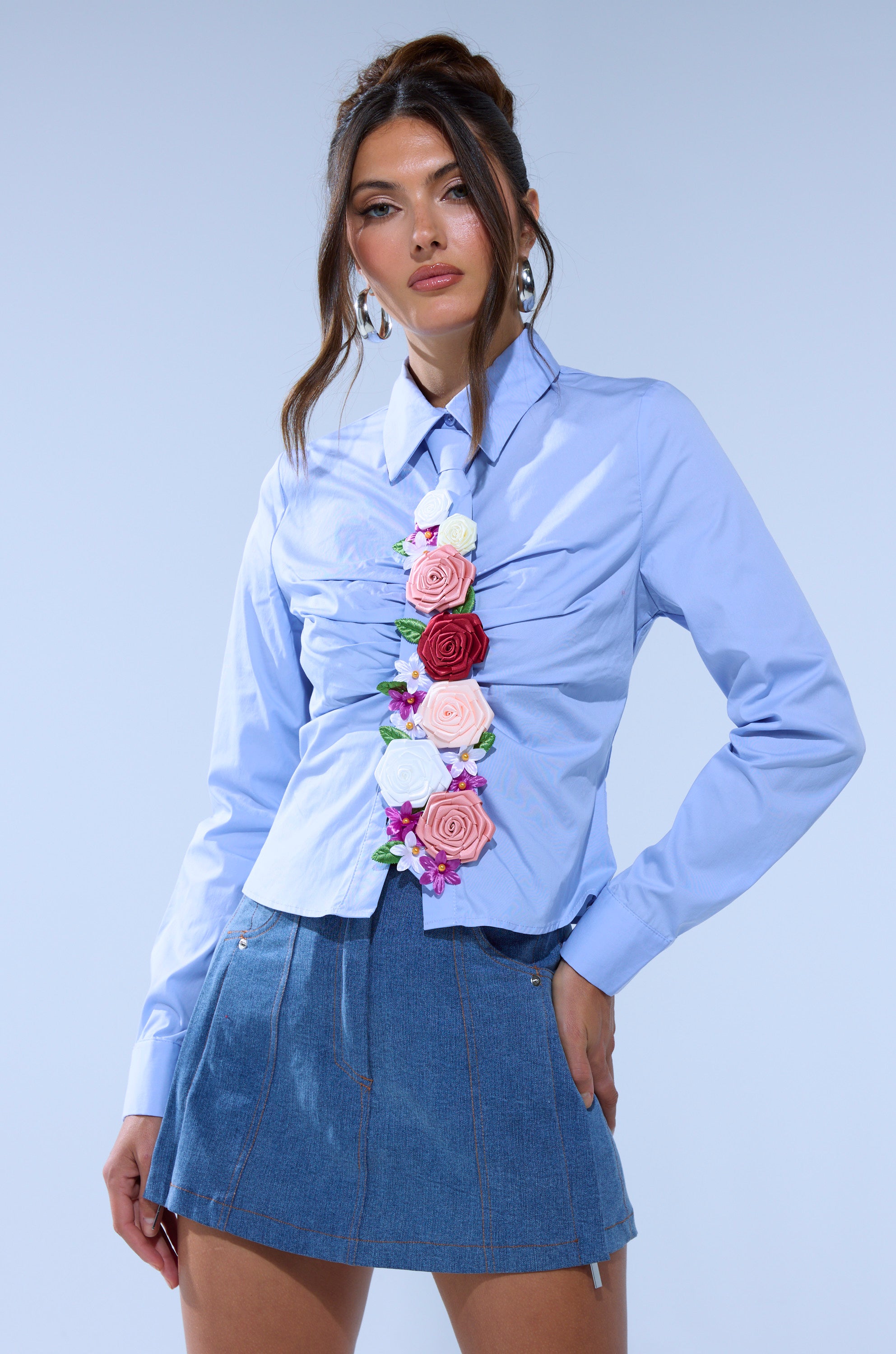 A woman models the BOUQUET BUTTON DOWN SHIRT with floral appliqués and a short denim skirt, pairing the look with statement earrings and a high updo with loose tendrils.