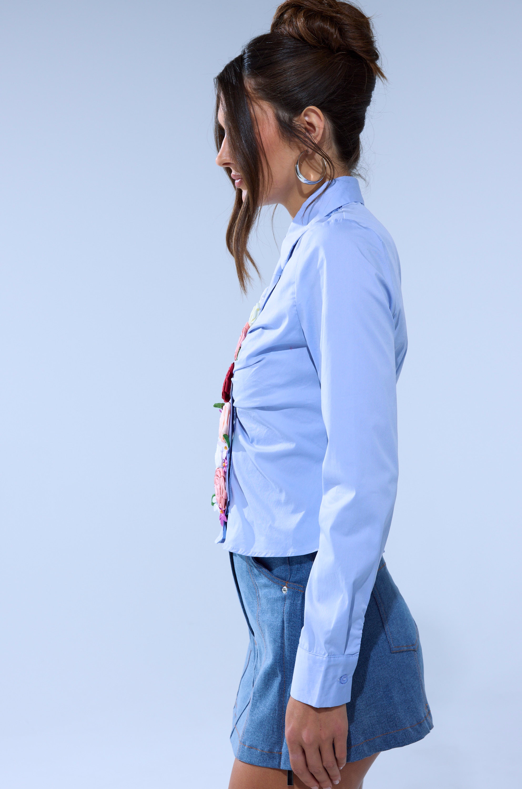 A woman with long brown hair in a bun, wearing large hoop earrings and the BOUQUET BUTTON DOWN SHIRT, pairs it with a short denim skirt while standing in profile against a plain light background.