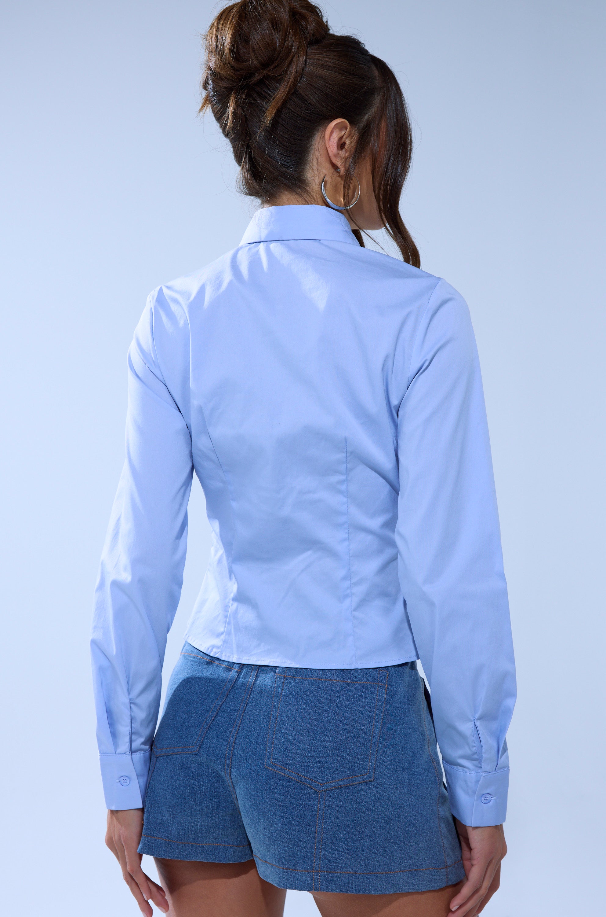 A woman with long brown hair in a bun is seen from the back, wearing the BOUQUET BUTTON DOWN SHIRT in light blue with long sleeves and blue denim shorts against a plain, light-colored background.