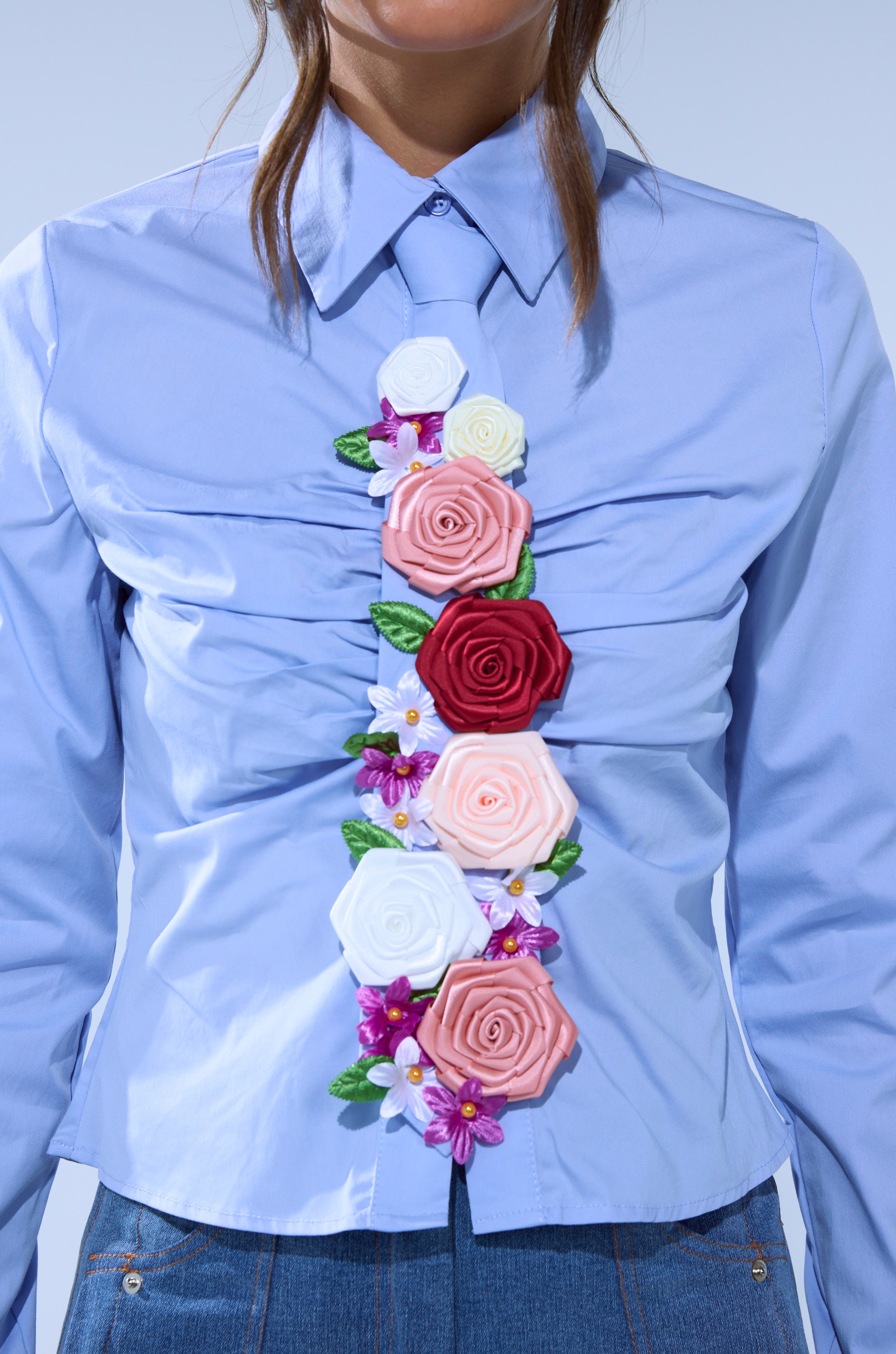 Someone wears the light blue BOUQUET BUTTON DOWN SHIRT with jeans, featuring a tie decorated with colorful floral appliques—red, pink, white, and purple—down the shirt's center.