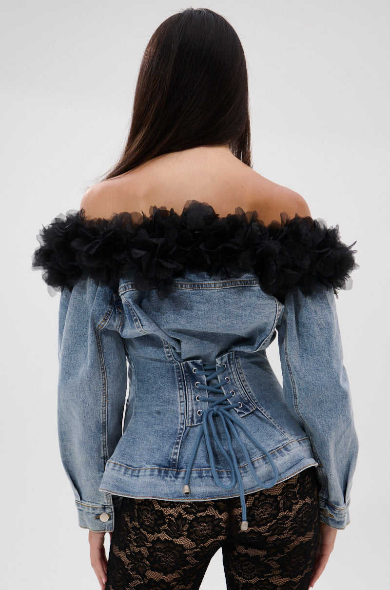 A woman with long dark hair is seen from behind, wearing the DANA OFF THE SHOULDER RUFFLE DETAIL DENIM TOP featuring a lace-up back and black ruffled trim, paired with black lace pants and floral tulle ruffle, against a plain light background.