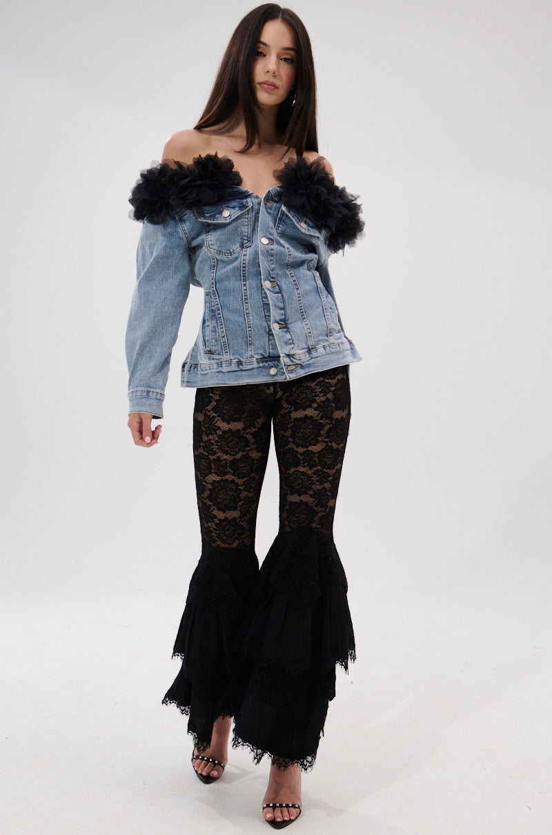 A woman in the DANA OFF THE SHOULDER RUFFLE DETAIL DENIM TOP, paired with black lace flared pants with ruffle hems and strappy heels, stands against a plain white background.