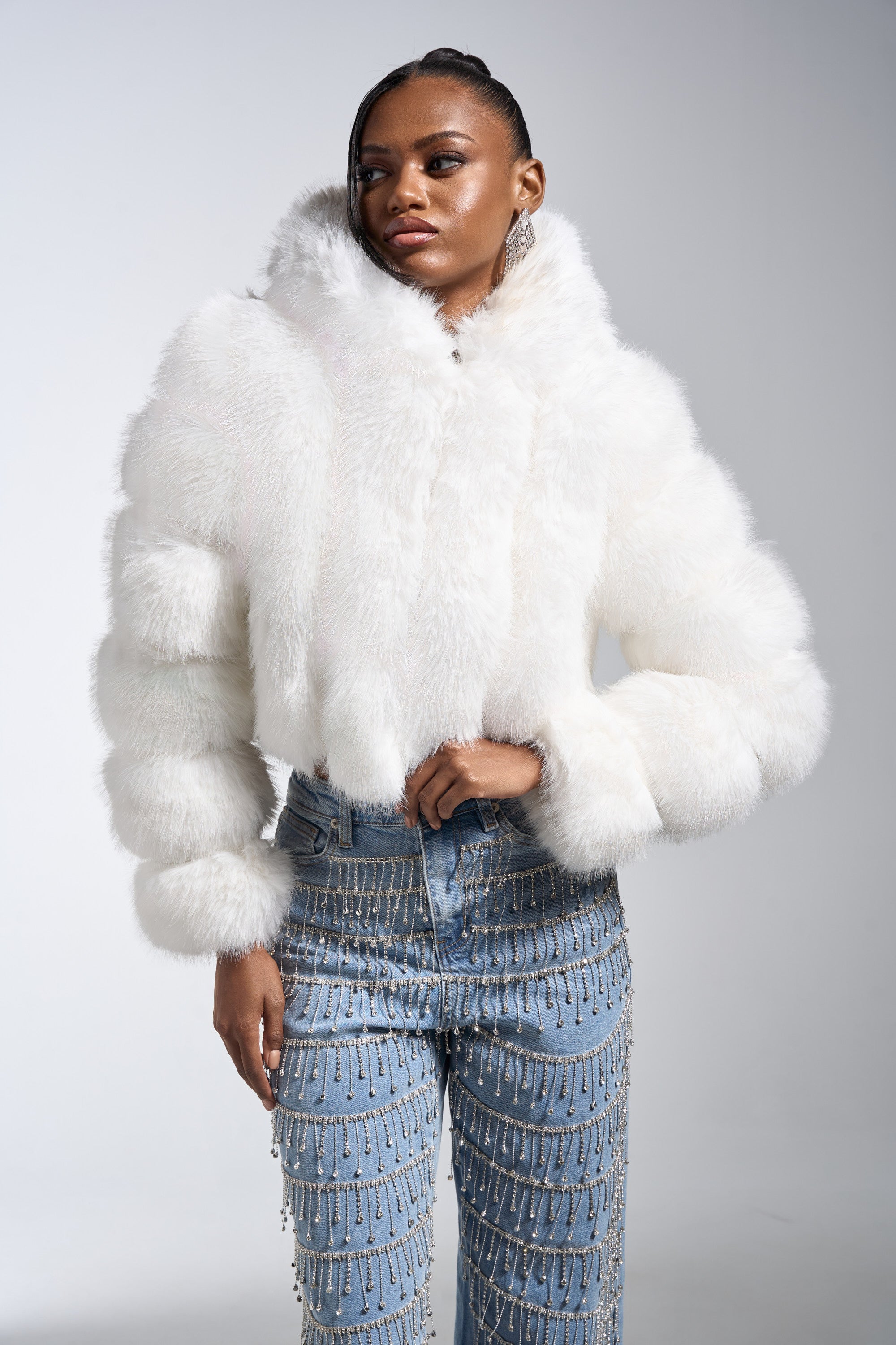 A woman wears the JOLENE FAUX FUR COAT IN WHITE with high-waisted blue jeans adorned with sparkling dangling chains, standing against a plain light gray background.