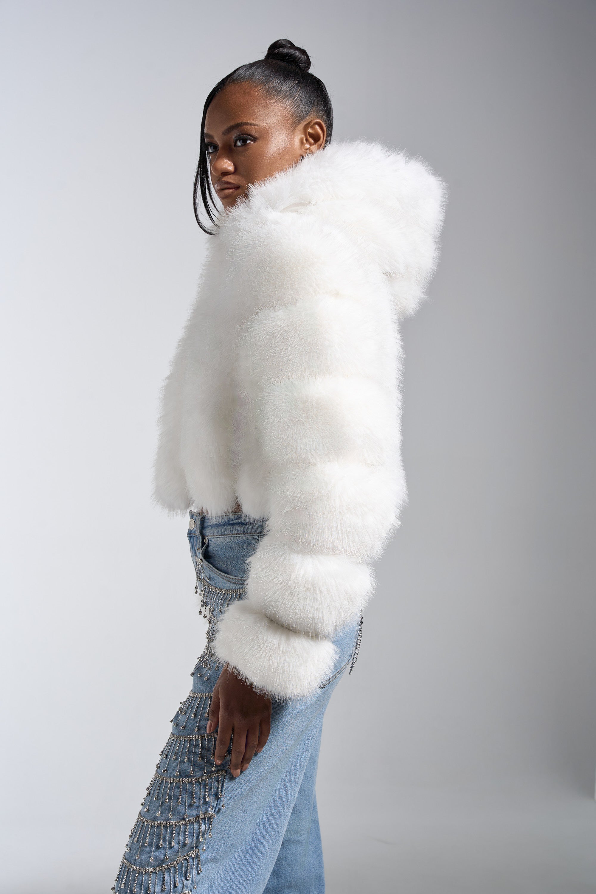 A woman models the JOLENE FAUX FUR COAT IN WHITE with an oversized hood, paired with light blue crystal fringe jeans, against a plain light gray background.