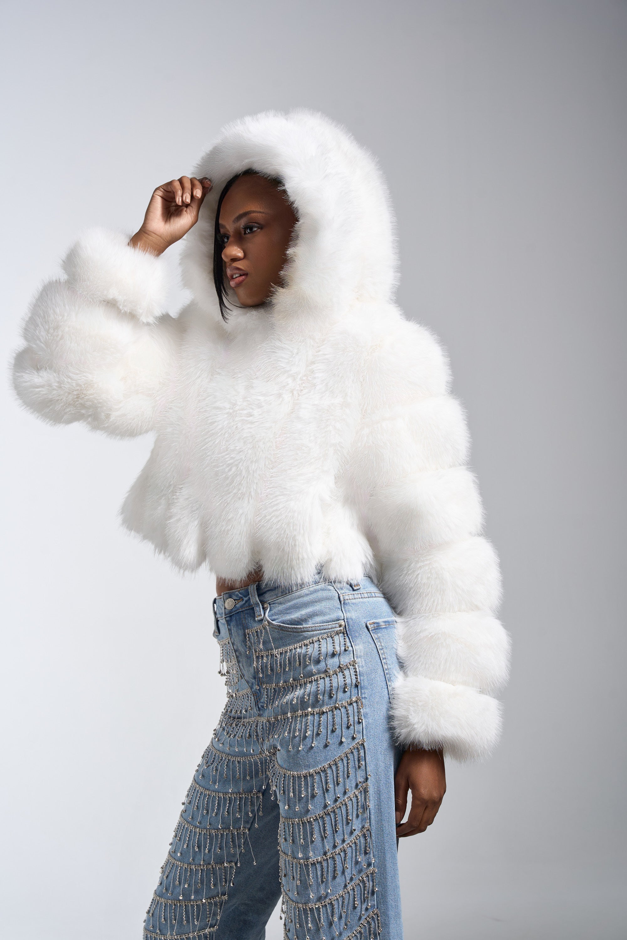 A woman poses in the JOLENE FAUX FUR COAT IN WHITE, featuring an oversized hood, paired with blue jeans adorned with silver chains, set against a plain light gray background.