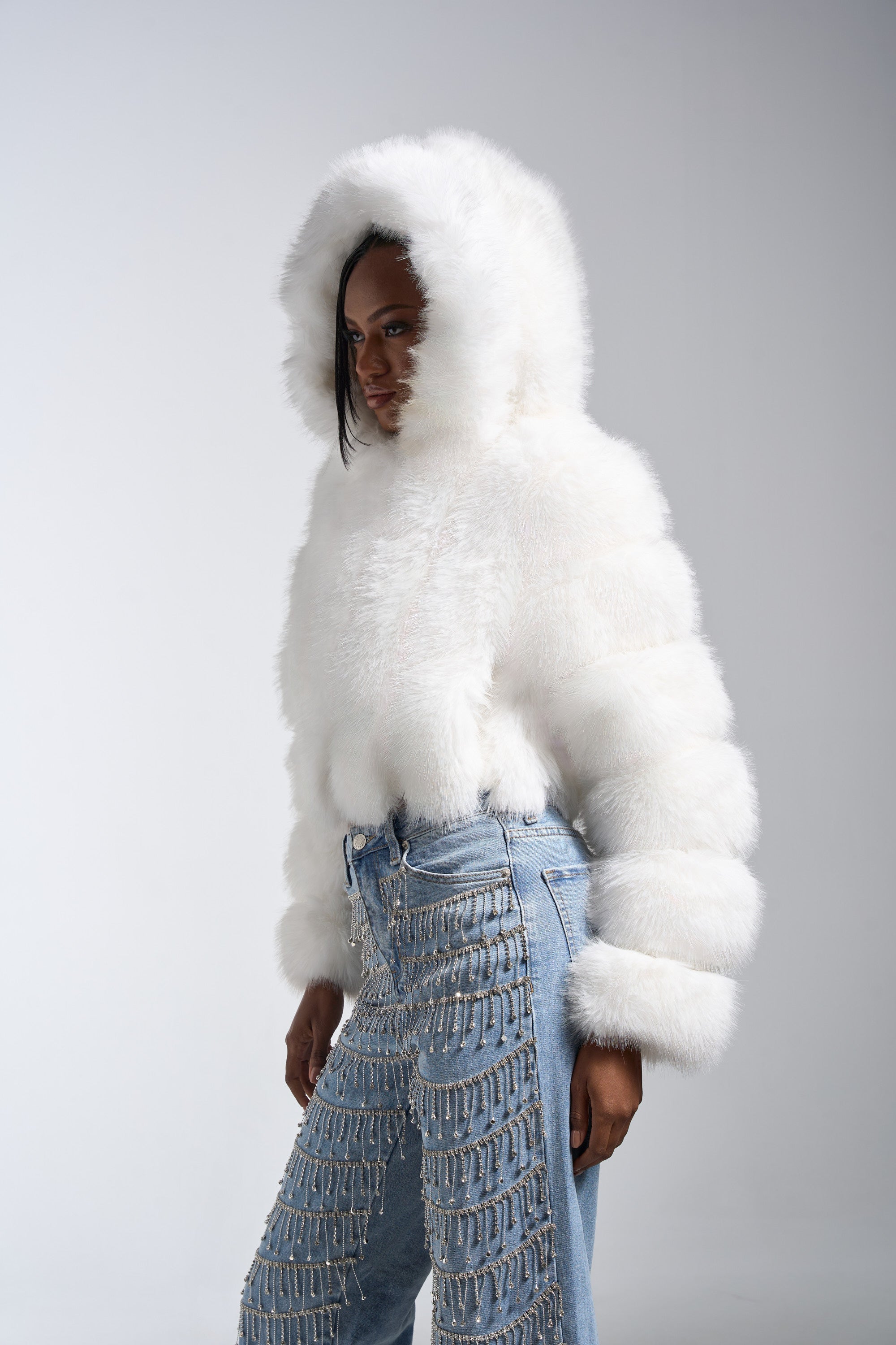 Someone models the JOLENE FAUX FUR COAT IN WHITE, featuring a hood, paired with high-waisted blue jeans with silver chains, standing against a plain light gray background.