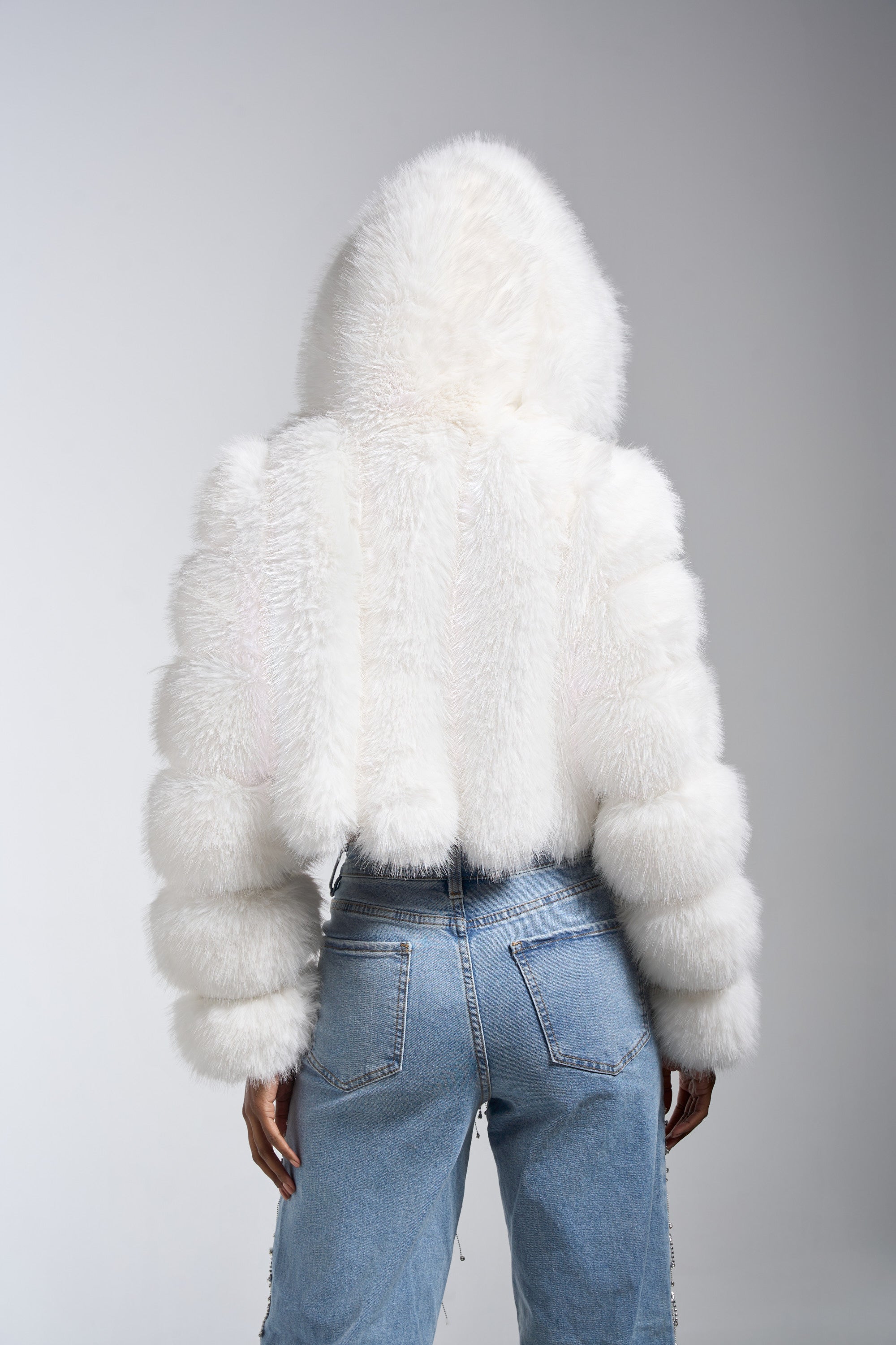 A person faces away, wearing the JOLENE FAUX FUR COAT IN WHITE with a hood and light blue high-waisted jeans. The plain background draws attention to the coat's fluffy texture and the denim.