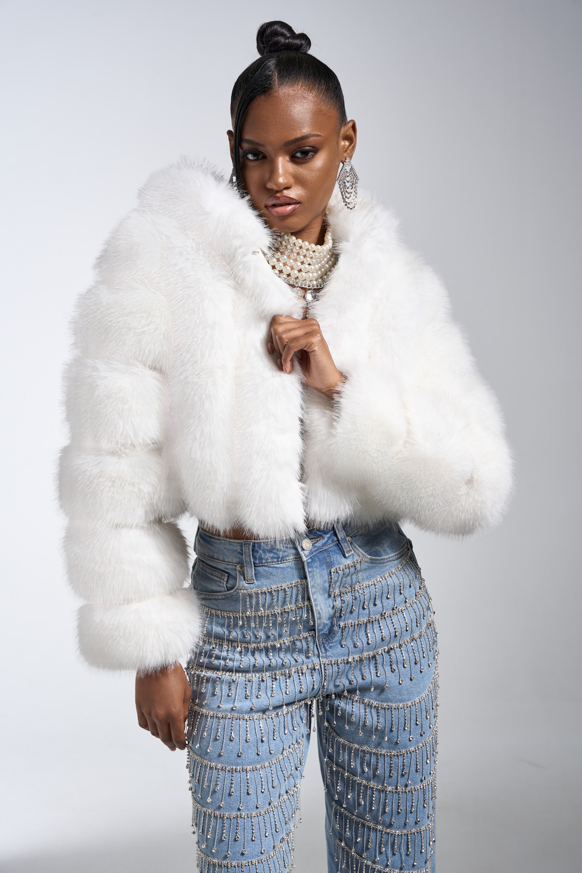 A woman poses in the JOLENE FAUX FUR COAT IN WHITE with an oversized hood, layered necklaces, statement earrings, and high-waisted jeans with dangling chains, set against a neutral background.