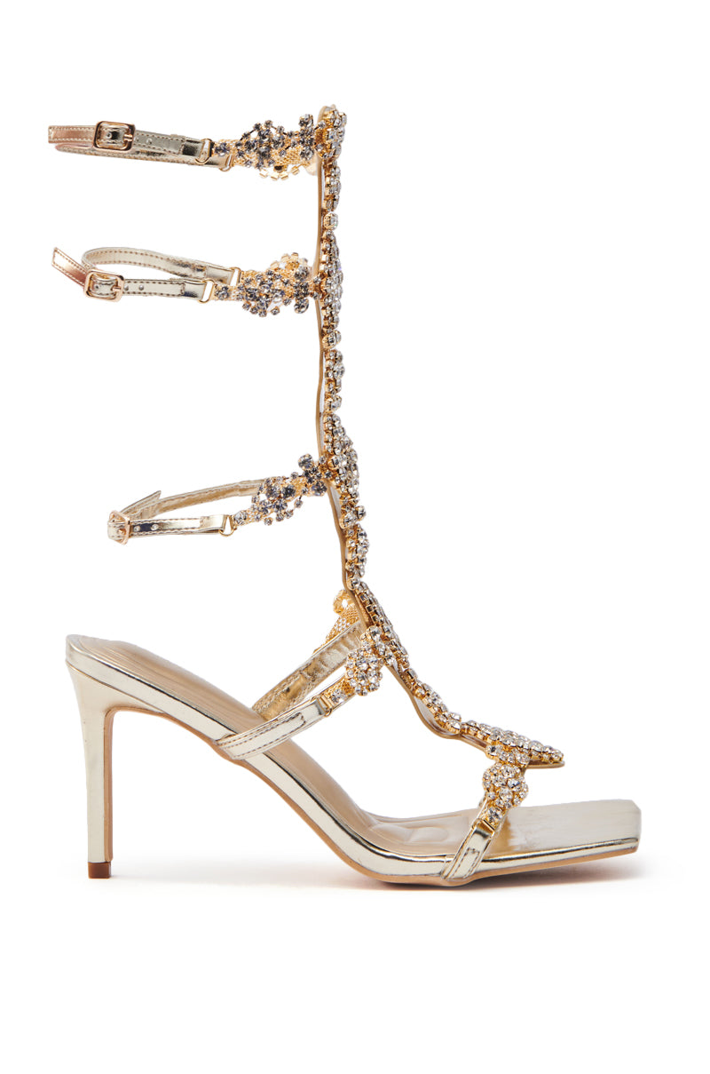 The AZALEA WANG JONQUIL GOLD EMBELLISHED GLADIATOR SANDAL is a strappy high heel with a metallic faux leather finish, featuring intricate beading, radiant rhinestone embellishments along the straps, and multiple buckles for elegant ankle wrapping.