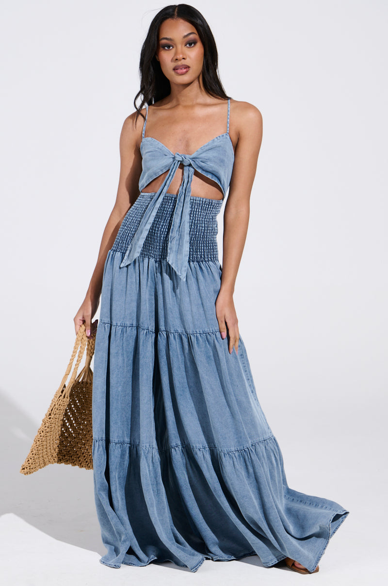 A woman poses against a plain backdrop in the "Jumpin Around Jumpsuit," featuring a blue sleeveless design with adjustable tank straps, a chest bow, and a smocked waist. She carries a woven handbag while her hair falls naturally.